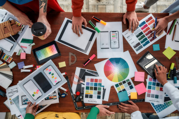 Working on a new project together. Top view of designers discussing sketches, choosing colors from palettes lying on the desk while having a meeting in the modern office