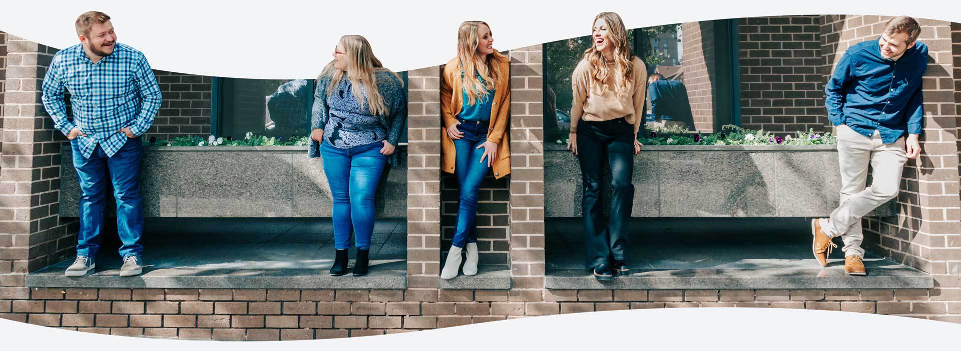 5 Metric Marketing team members stand and lean against brick and stone architectural features, smiling and engaging with each other